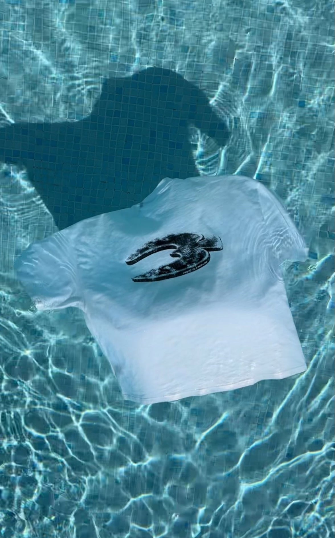 A white short-sleeve T-shirt with a black dove graphic on the back, floating on clear blue pool water with sunlight and ripples.