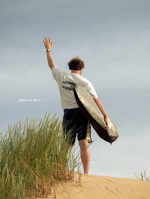 A man standing on a sandy dune with sea oats, seen from behind, holding a surfboard under his arm and waving his other hand toward the sky. He is wearing a white t-shirt and dark shorts.